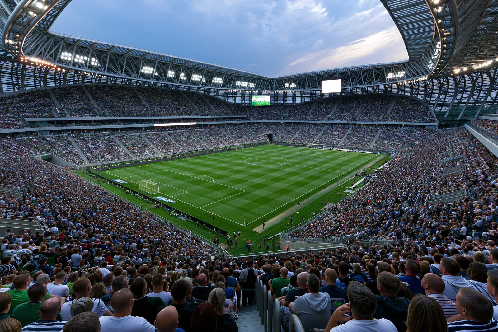 Didelis futbolo stadionas, vasaros vakarą apšviestas prožektoriais, pilnas sirgalių.