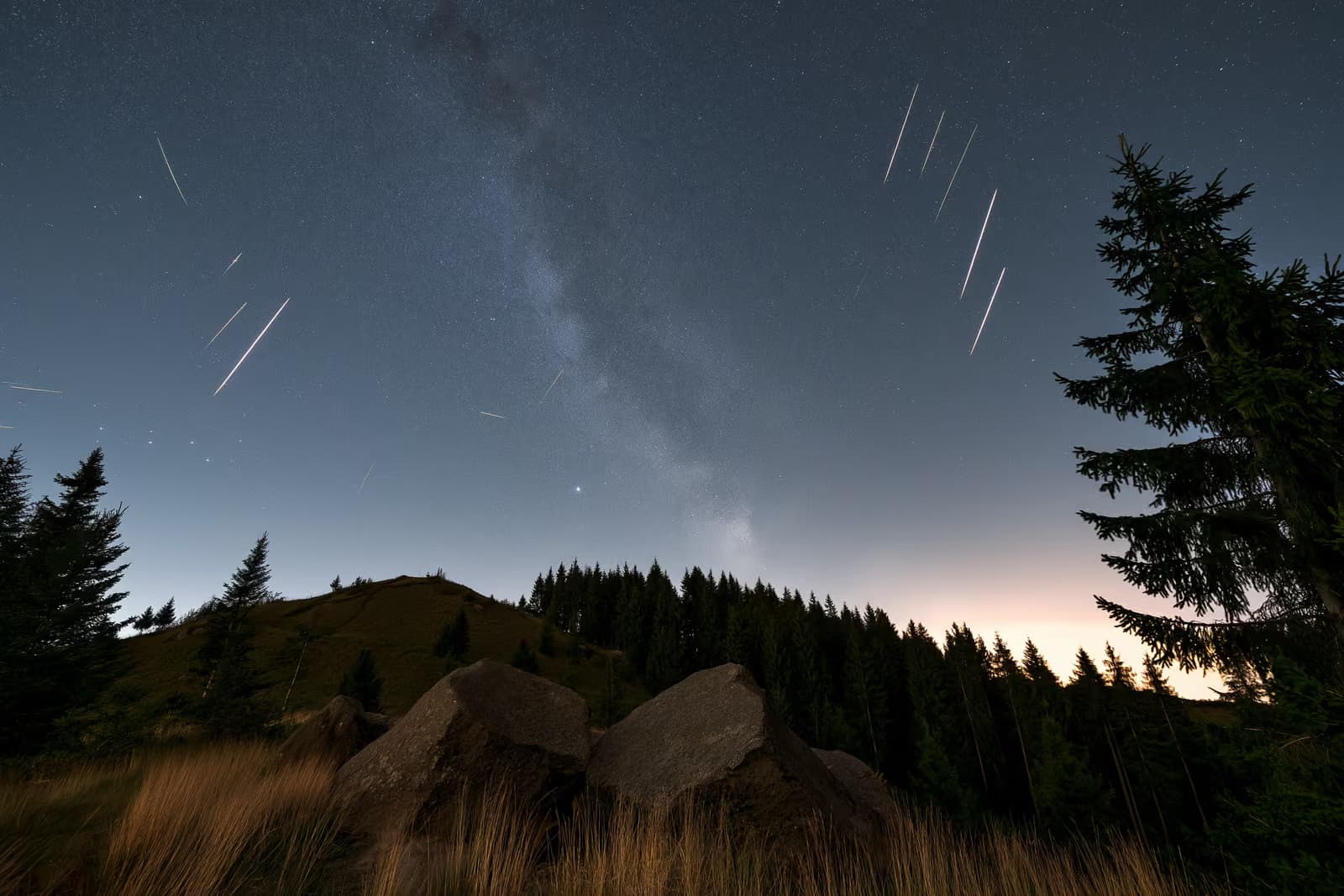 Naktinis dangus virš tamsaus Vokietijos kaimo su meteorų ruožais ir blankia žvaigždžių juosta viršuje.